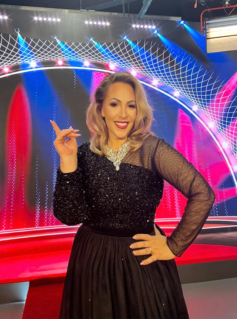Smiling woman in a black sequined top and sheer sleeves posing on a colorful stage with neon lights and a red floor background.