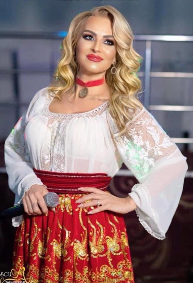 Woman in a white lace blouse and red embroidered skirt, holding a microphone, posing on stage.